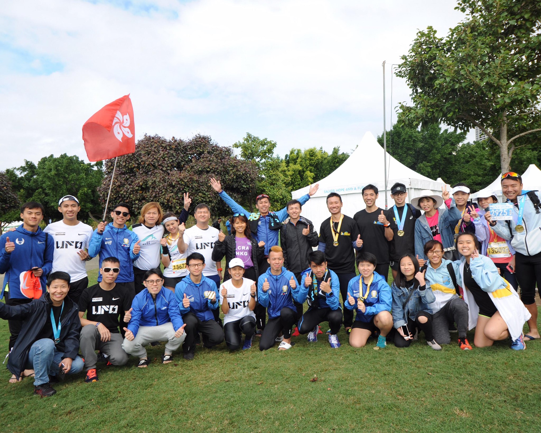 A group photo of the Ekiden Sports Management team at an outdoor event. Team members are wearing branded sports apparel in blue and white, posing together on a grass field with the Hong Kong flag, showcasing a professional and energetic team culture.