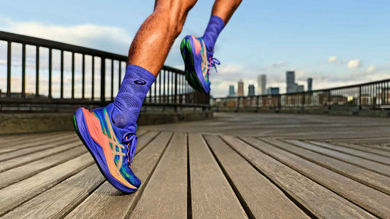 A runner in vibrant purple performance gear runs along a wooden boardwalk in a city park at dawn. The runner wears bold, patterned purple crew socks with a black logo, and technically advanced blue and purple running shoes with striking neon orange and green accents. A detailed low-angle perspective focuses on the powerful mid-stride of the runner’s legs. The background features a dark railing and a distant, misty city skyline under a soft, golden and blue sky. The scene conveys a sense of dynamic energy, athletic discipline, and a quiet, determined morning run.